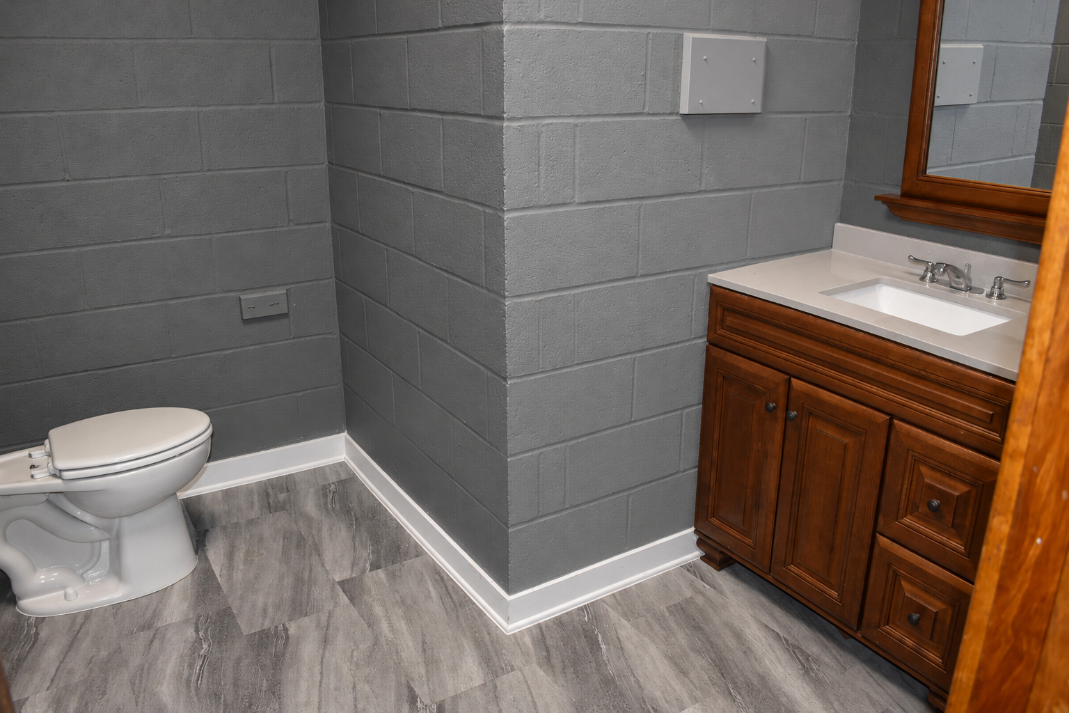 Wide gray-block restroom with a closed white toilet on the left, central partition, brown wood vanity, clear white countertop and mirror on the right