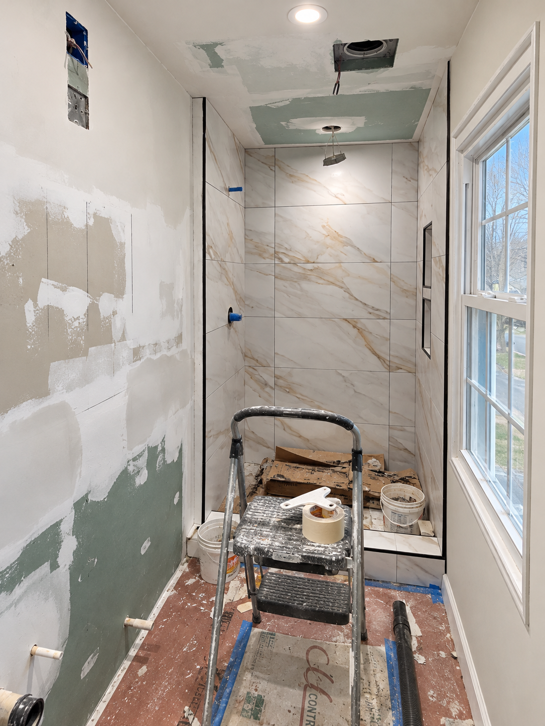 Bathroom rebuild with unfinished wall and ceiling surfaces, marble-look shower tiles, a stepladder, work materials and floor protection.