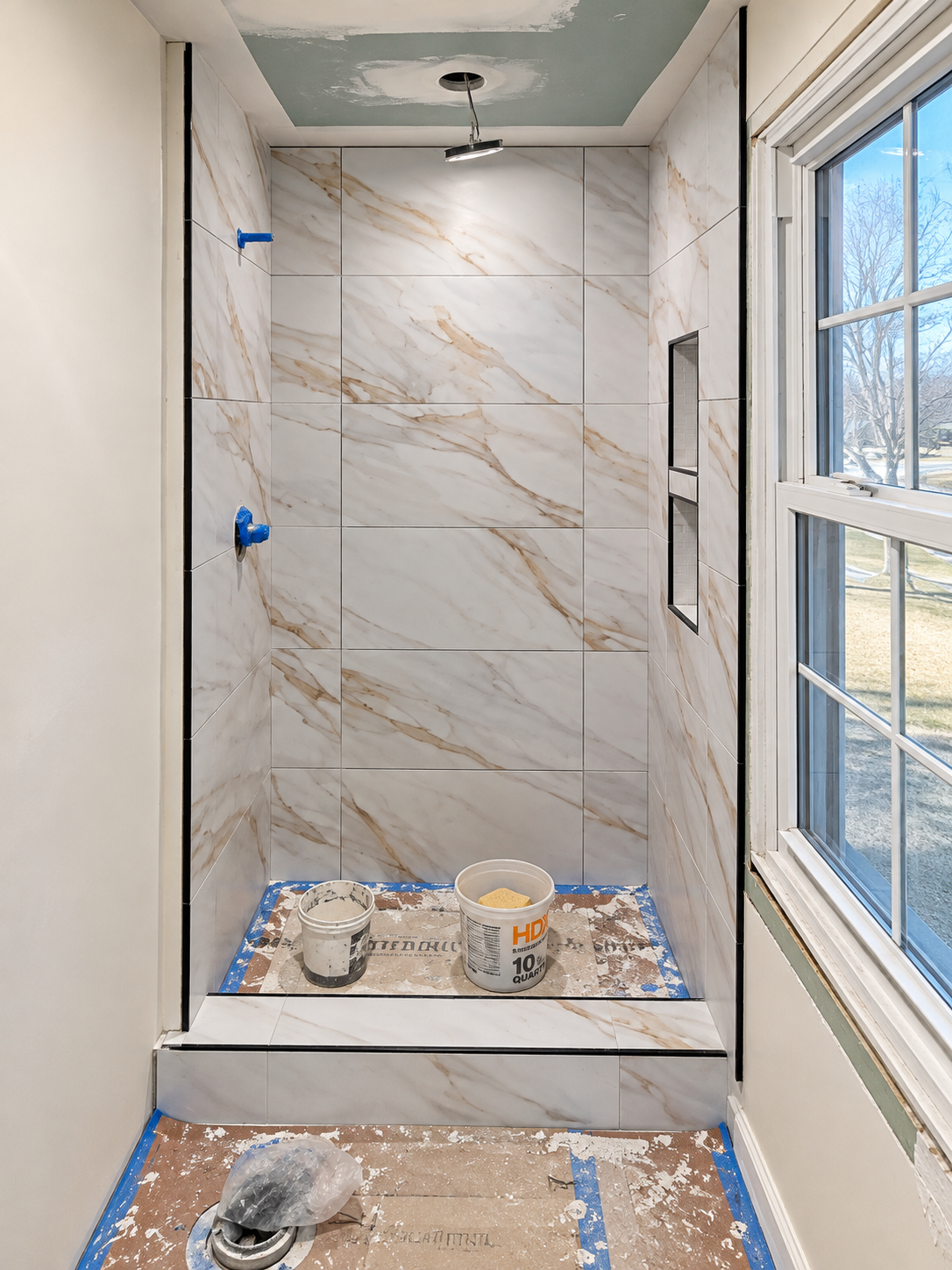 Marble-look shower wall tiles with two vertically stacked recessed niches, black edging, an unfinished ceiling opening and buckets on floor protection.