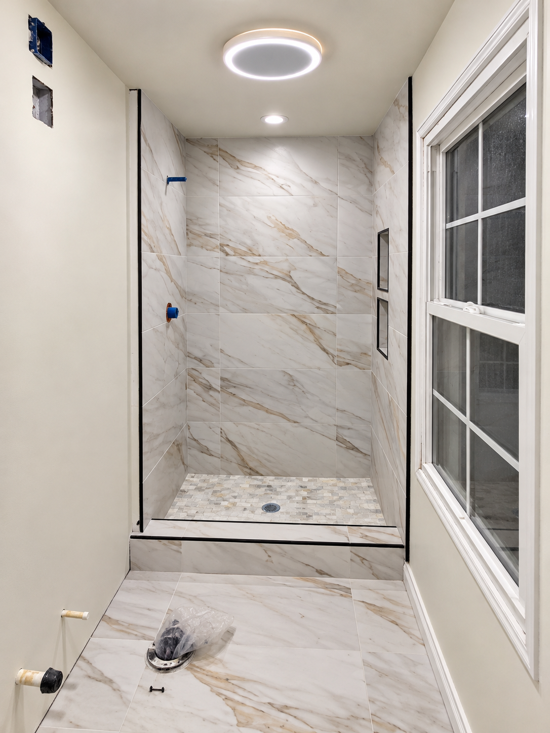 Bathroom before fixture and shower-door installation with mosaic shower flooring, a tiled curb, wall niches and marble-look floor and wall tiles.