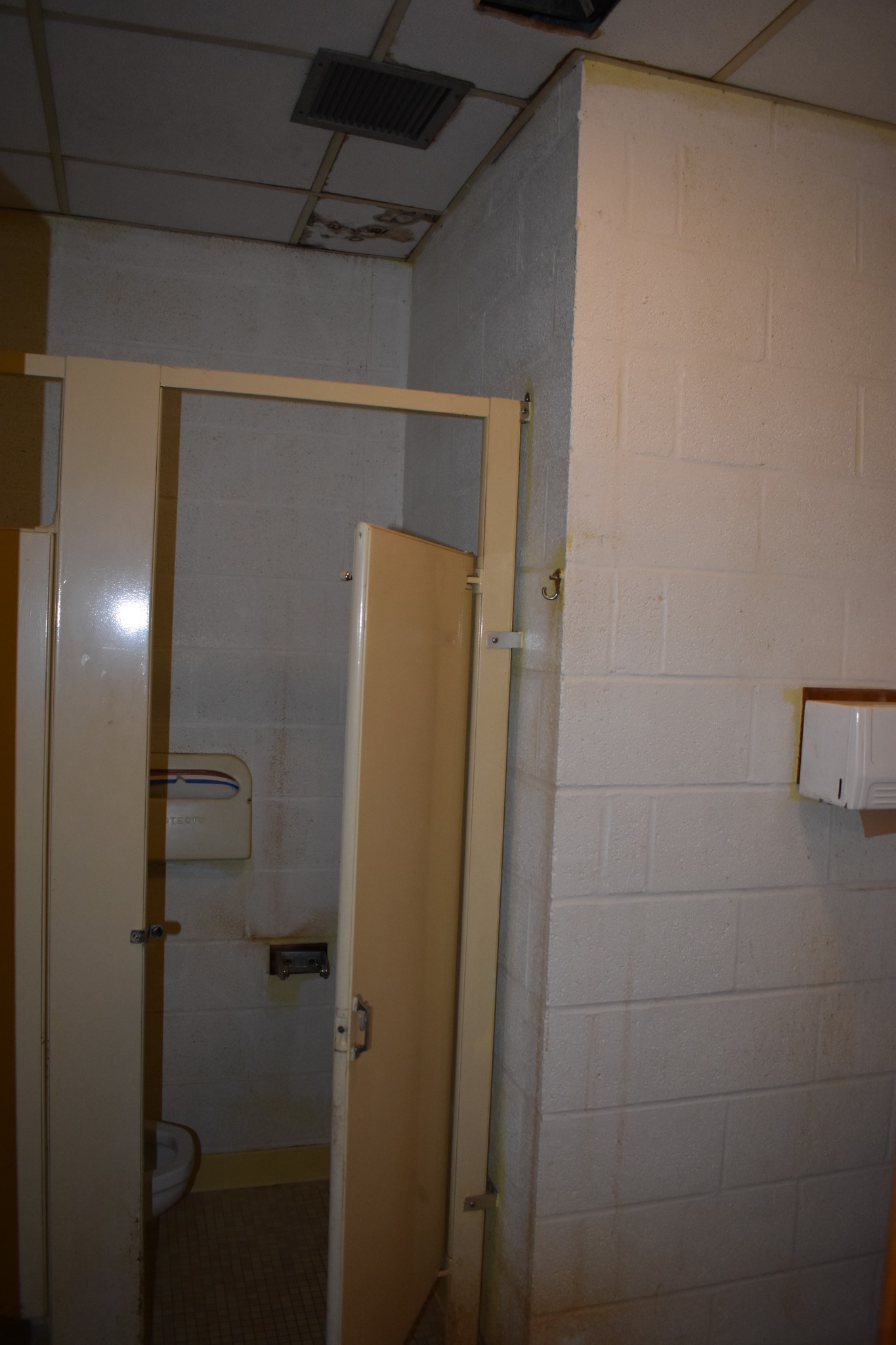 Restroom with an open beige stall door at left and center, white block partition on the right and stained ceiling tiles above