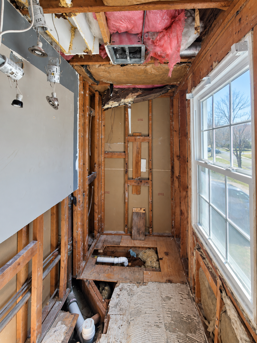 Bathroom during demolition with exposed wood framing, insulation, ceiling fixtures, floor openings, drain piping and a window.
