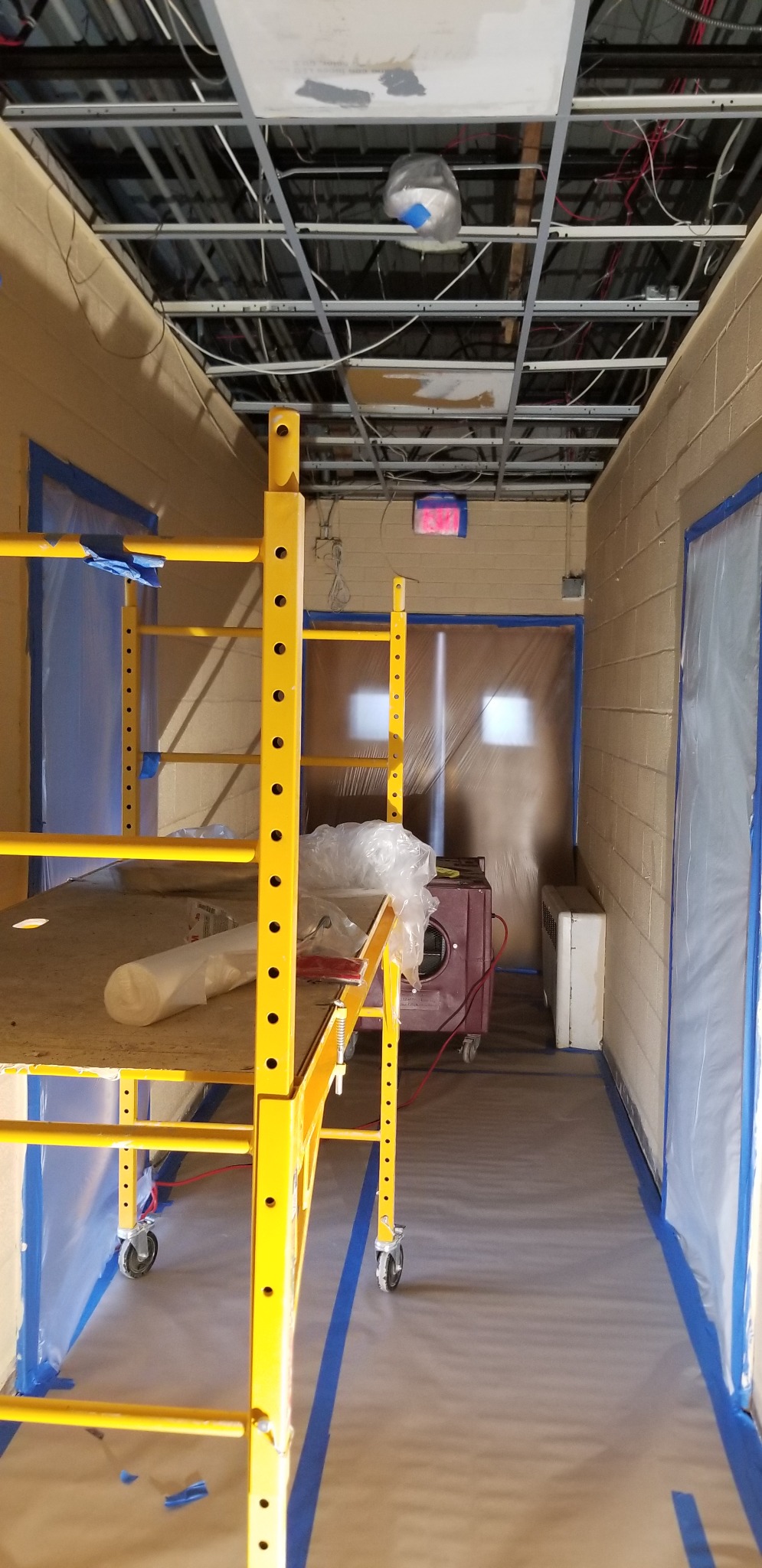 Rolling scaffold beneath the exposed hallway ceiling grid with protected doorways and flooring