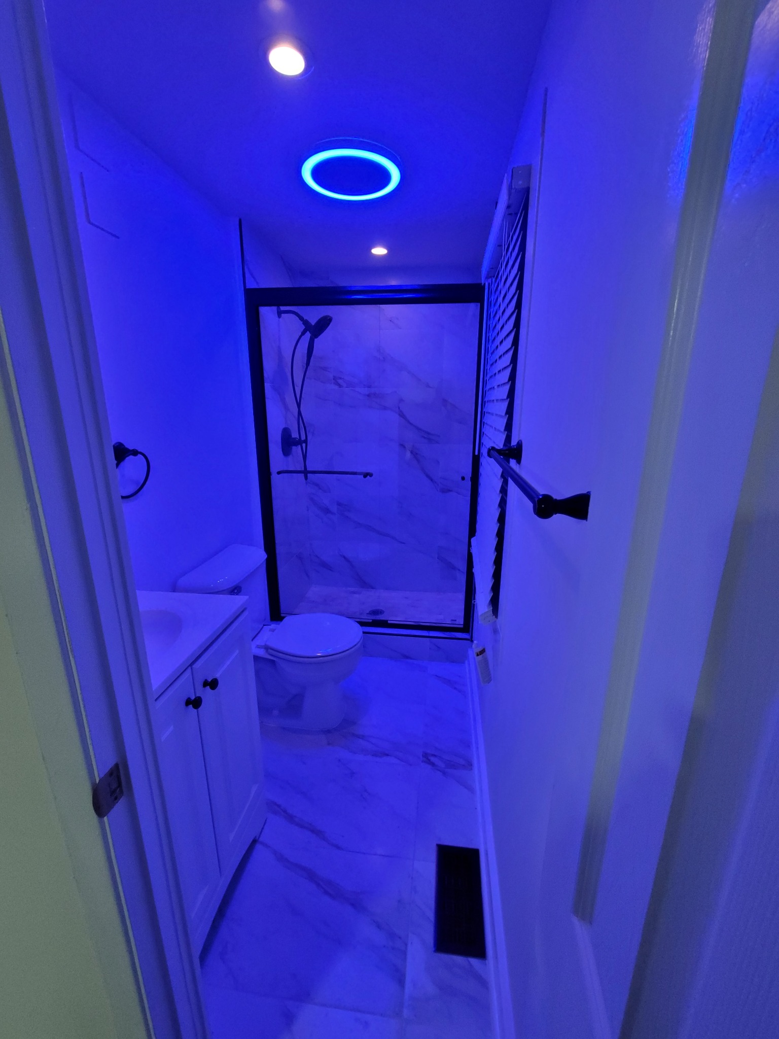 Finished bathroom viewed from the doorway with blue accent lighting, white ceiling lights, a black-framed glass shower enclosure, white vanity, toilet, tiled floor and black towel bar.