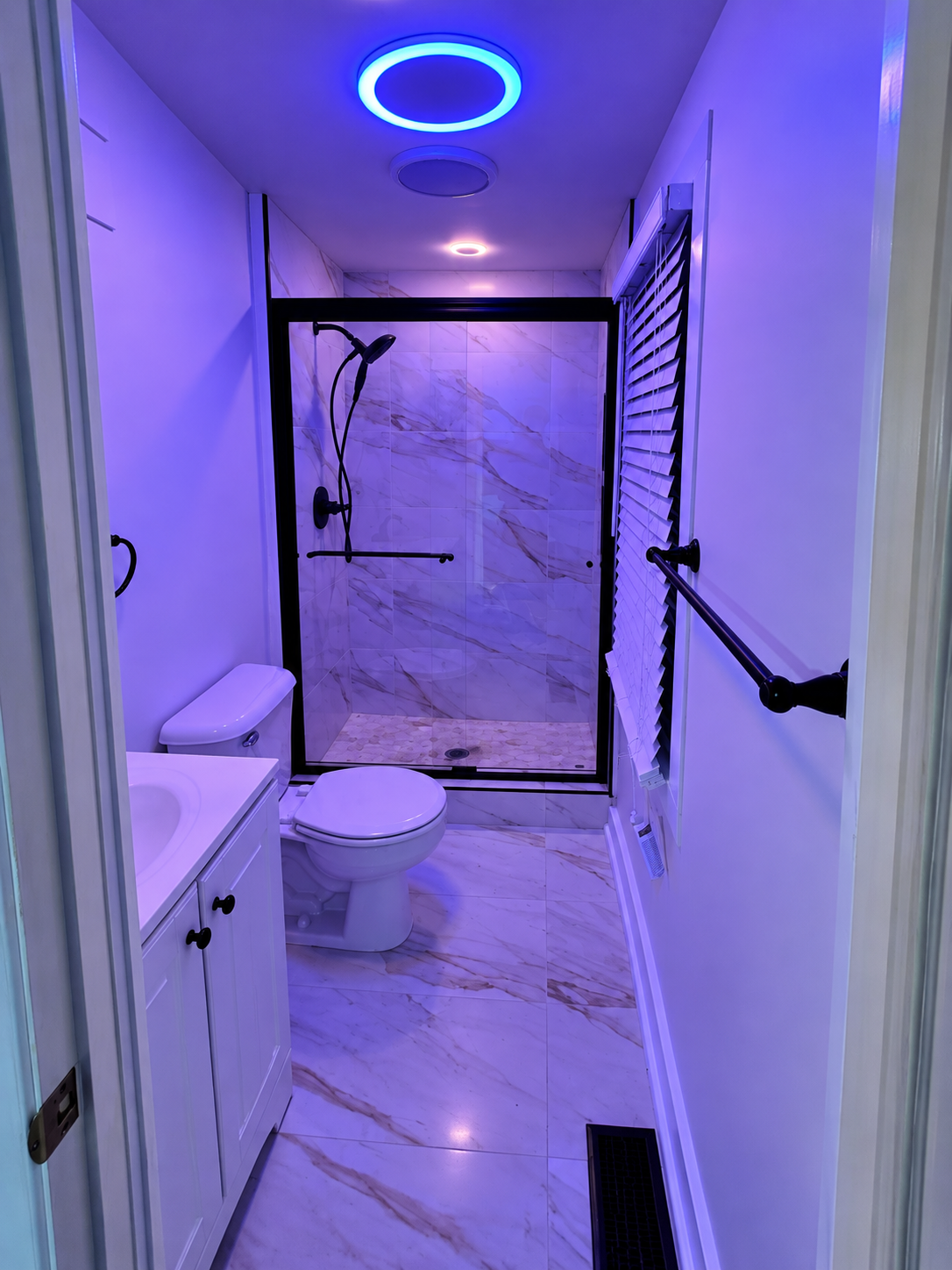 Finished bathroom presentation with blue-purple ceiling accent lighting, black-framed glass shower enclosure, white vanity, toilet and tiled floor.
