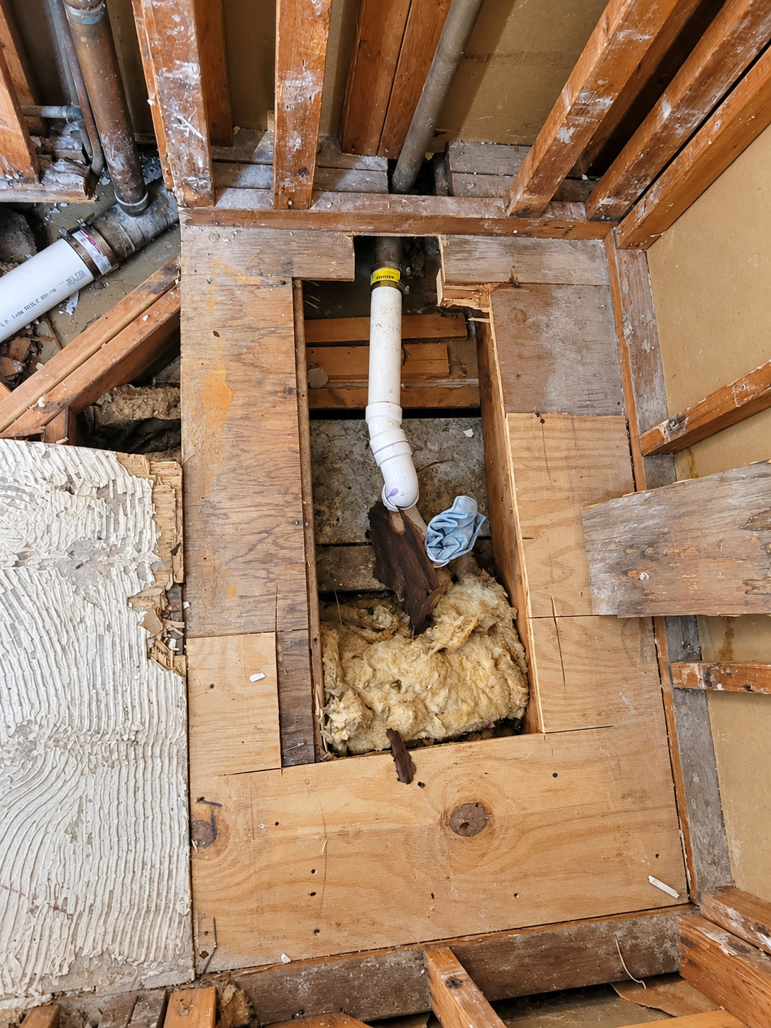 Close view of opened wood subfloor, white drain piping, insulation and adjacent exposed wall framing.