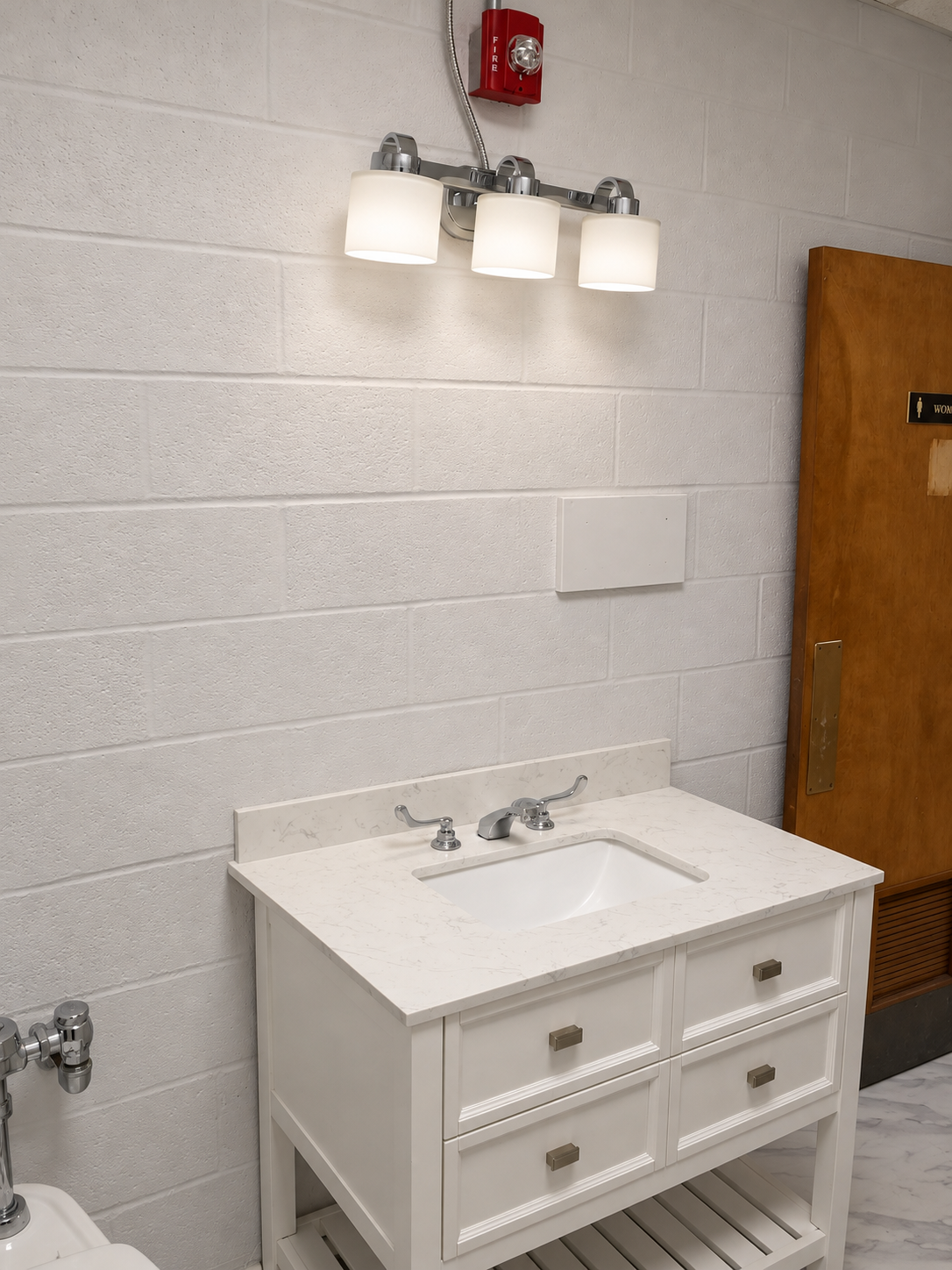 White open-shelf vanity with pale countertop, chrome faucet, three-shade light, light block wall, red alarm and wood door