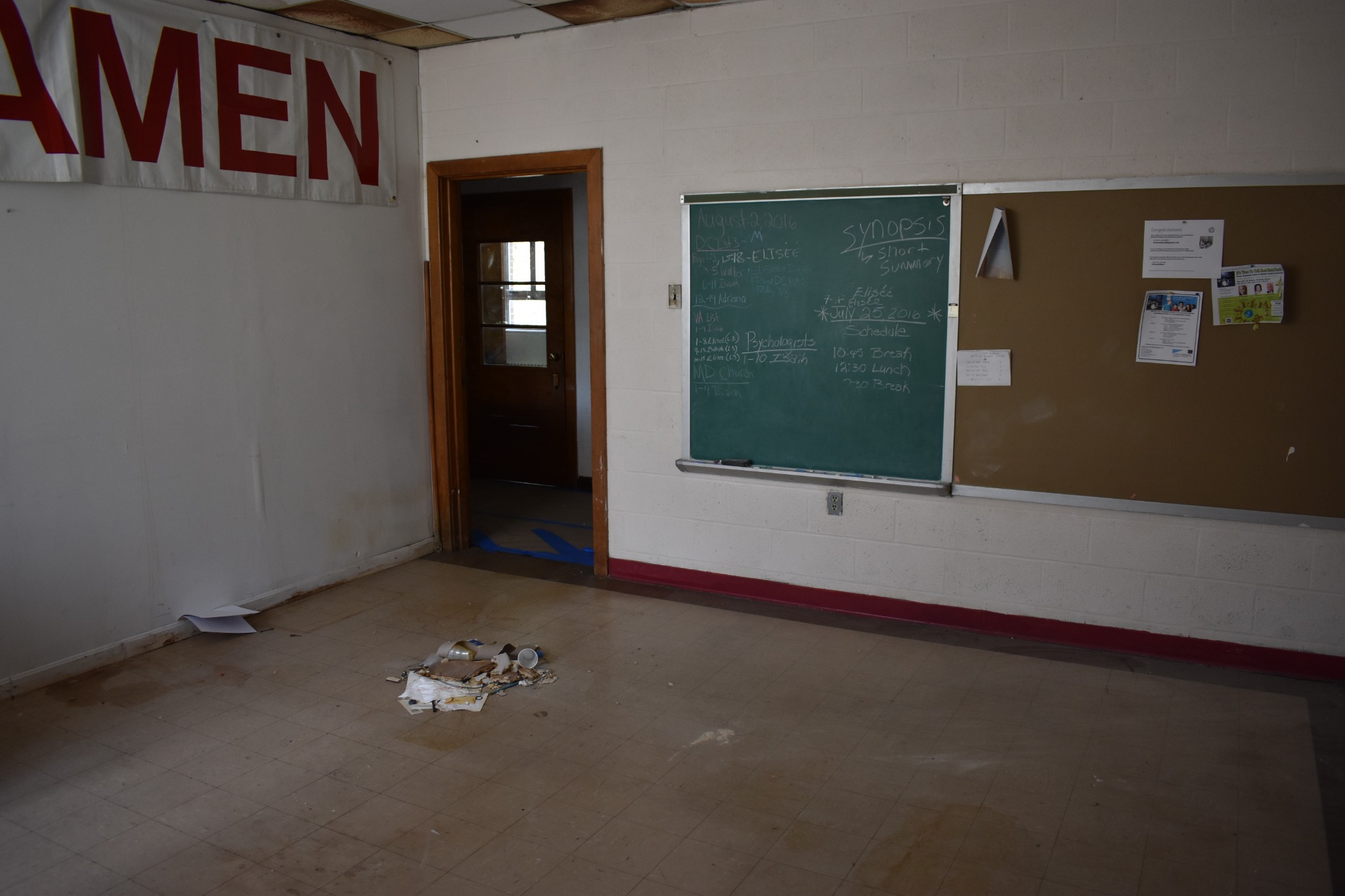 Classroom before restoration with an AMEN banner, doorway, chalkboard, bulletin board and floor debris
